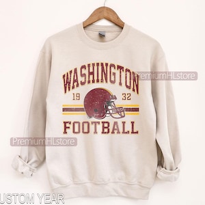 May include: A beige crewneck sweatshirt featuring a distressed graphic with the text WASHINGTON FOOTBALL, the numbers 19 and 32, and a maroon football helmet illustration centered between two horizontal stripes in maroon and yellow colors.
