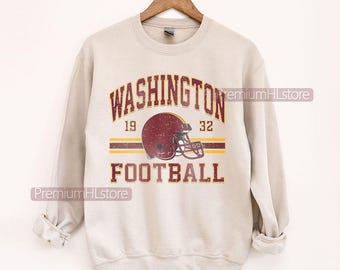Op de afbeelding: Een beige sweater met ronde hals voorzien van een verweerde print met de tekst WASHINGTON FOOTBALL, de getallen 19 en 32, en een illustratie van een bordeauxrode footballhelm gecentreerd tussen twee horizontale strepen in bordeaux en geel.