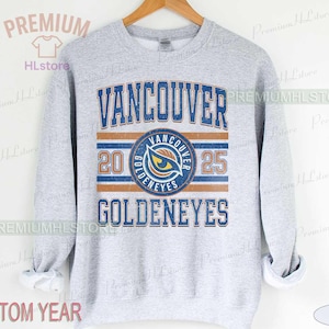 May include: A grey sweatshirt featuring the text "VANCOUVER GOLDENEYES" in blue and orange. The design includes a circular emblem with an eye graphic and the years "20" and "25". The sweatshirt is displayed on a wooden hanger.