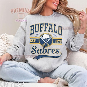 May include: A light gray sweatshirt featuring the text "BUFFALO Sabres" in navy blue and gold, with a logo of crossed swords and a bison. The sweatshirt includes the text "EST. 1970".