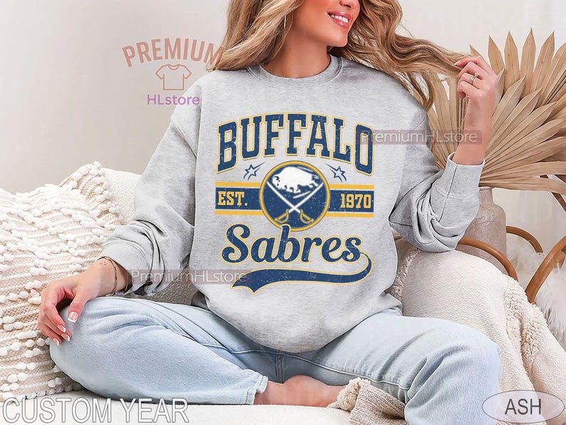 May include: A light gray sweatshirt featuring the text "BUFFALO Sabres" in navy blue and gold, with a logo of crossed swords and a bison. The sweatshirt includes the text "EST. 1970".