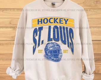 May include: A cream-colored sweatshirt with a blue and yellow graphic design. The graphic features the text "HOCKEY ST. LOUIS EST 1967" and a hockey helmet.