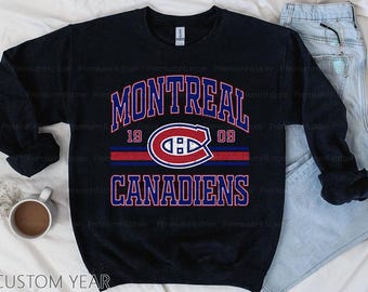 Puede incluir: Una sudadera azul marino con el texto "MONTREAL CANADIENS" en una fuente universitaria. El diseño incluye el logotipo del equipo y el año "1909" en rojo y azul. La sudadera se muestra sobre una superficie blanca con unos vaqueros y una taza de café.