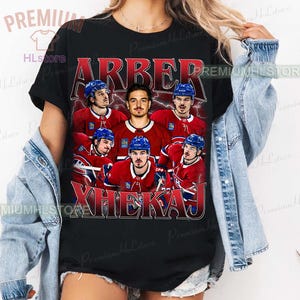 May include: Black t-shirt featuring a vintage-style graphic of a hockey team. The design includes the names "ARBER" and "XHEKAJ" in red and white lettering, along with images of the players in their red and blue uniforms.
