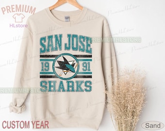 May include: A sand-coloured sweatshirt with the text "SAN JOSE SHARKS" in teal, the years "19" and "91", and a shark logo. The sweatshirt is displayed on a wooden hanger.