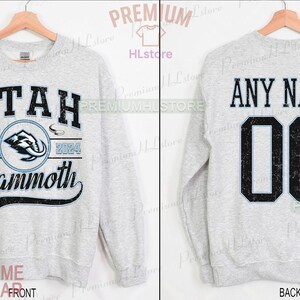 K&ouml;nnte beinhalten: Hellgraues Sweatshirt mit dem Text "UTAH Mammoth" und einem Mammut-Logo auf der Vorderseite. Auf der R&uuml;ckseite stehen "ANY NAME" und die Nummer "00". Das Sweatshirt ist aschfarben.