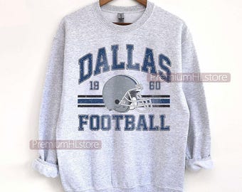 Op de afbeelding: Een lichtgrijs sweatshirt met het woord "DALLAS" in blauwe blokletters boven een grijze footballhelm-afbeelding. De tekst "FOOTBALL" staat onder de helm. Het jaartal "1960" is boven de helm gedrukt.