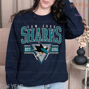 May include: Navy blue sweatshirt featuring the San Jose Sharks logo in teal and white, with the text "SAN JOSE SHARKS EST. 1991". The sweatshirt has a crew neck and long sleeves. The text "CUSTOM YEAR" is printed at the bottom.