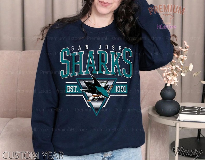 May include: Navy blue sweatshirt featuring the San Jose Sharks logo in teal and white, with the text "SAN JOSE SHARKS EST. 1991". The sweatshirt has a crew neck and long sleeves. The text "CUSTOM YEAR" is printed at the bottom.