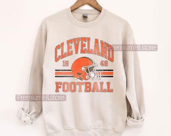 May include: A light beige sweatshirt featuring a distressed orange and brown graphic. The graphic reads "CLEVELAND FOOTBALL" with a football helmet. The sweatshirt is hanging on a wooden hanger.