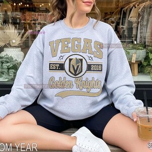 Puede incluir: Una sudadera gris claro con el texto "VEGAS Golden Knights" en dorado y negro. La sudadera tiene cuello redondo y mangas largas. Tambi&eacute;n es visible el texto "EST. 2016". Tambi&eacute;n son visibles las palabras "CUSTOM YEAR" y "SPORT GREY".
