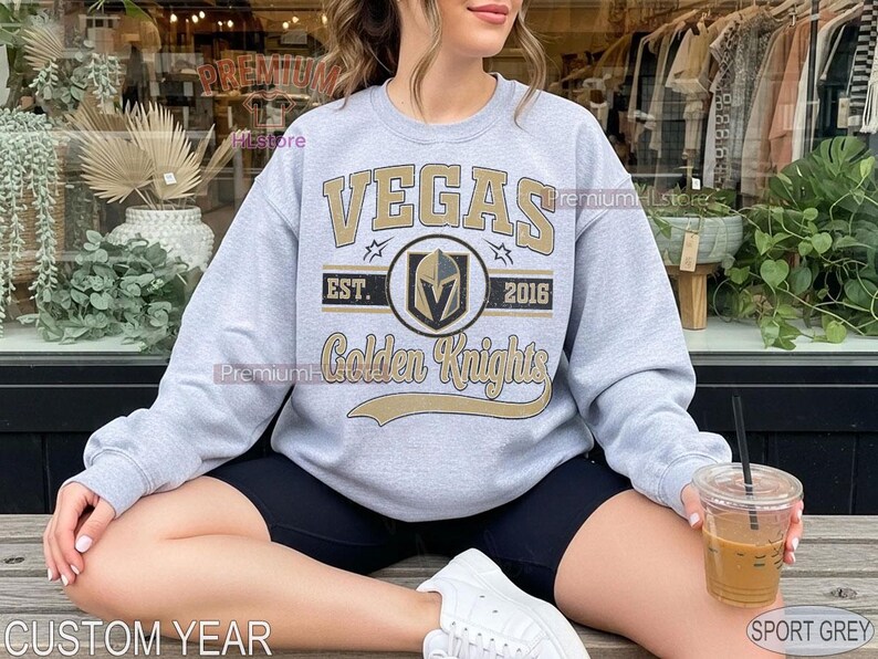 Puede incluir: Una sudadera gris claro con el texto "VEGAS Golden Knights" en dorado y negro. La sudadera tiene cuello redondo y mangas largas. Tambi&eacute;n es visible el texto "EST. 2016". Tambi&eacute;n son visibles las palabras "CUSTOM YEAR" y "SPORT GREY".