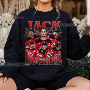 May include: Black sweatshirt featuring a graphic of a hockey player and the text "JACK HUGHES" in red. The design includes images of the player in a red and black jersey. The sweatshirt is a casual style.