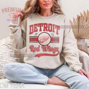 Puede incluir: Sudadera de color arena con el texto "DETROIT Red Wings" en rojo, con un logotipo de una rueda alada. El dise&ntilde;o incluye el a&ntilde;o de establecimiento "1926". La sudadera es de estilo casual.