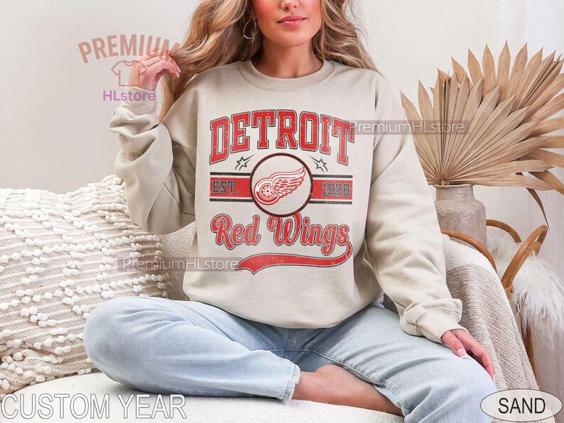 Puede incluir: Sudadera de color arena con el texto "DETROIT Red Wings" en rojo, con un logotipo de una rueda alada. El dise&ntilde;o incluye el a&ntilde;o de establecimiento "1926". La sudadera es de estilo casual.