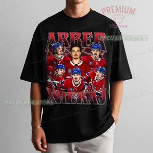 May include: Black t-shirt featuring a vintage-style graphic of a hockey team. The design includes the team's name in red, along with images of the players in their red and blue uniforms. The text "ARBER XHEKAJ" is prominently displayed.