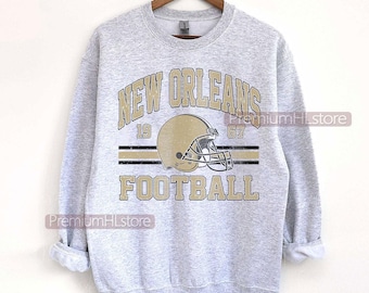 May include: A light gray sweatshirt with a vintage-style graphic. The design features the words "NEW ORLEANS FOOTBALL" in gold lettering, along with a gold football helmet and the numbers "19" and "67".