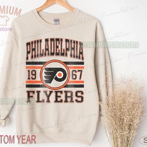Può includere: Una felpa color sabbia con la scritta "PHILADELPHIA FLYERS" in nero e arancione, il logo della squadra e i numeri "1967". La felpa ha un aspetto vintage e usurato.