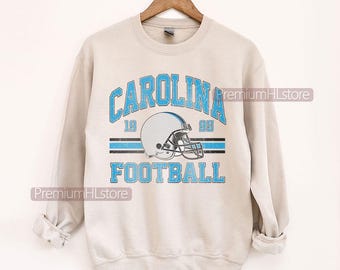 Może przedstawiać: Jasnoszara bluza z napisem "CAROLINA FOOTBALL" w jasnoniebieskim kolorze i grafiką kasku do futbolu amerykańskiego. Projekt zawiera liczby "19" i "95". Bluza wisi na drewnianym wieszaku.