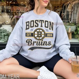 Puede incluir: Una sudadera gris claro con el texto "BOSTON Bruins" en negro y dorado. La sudadera presenta el logotipo de los Bruins y el a&ntilde;o "1924". La sudadera es de color gris deportivo.
