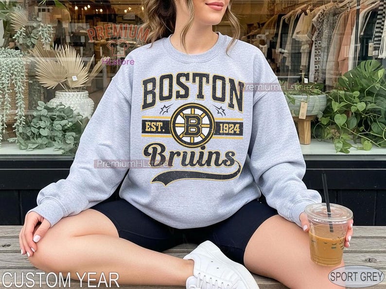Puede incluir: Una sudadera gris claro con el texto "BOSTON Bruins" en negro y dorado. La sudadera presenta el logotipo de los Bruins y el a&ntilde;o "1924". La sudadera es de color gris deportivo.