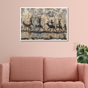 Pu&ograve; includere: Una stampa incorniciata raffigurante arte rupestre antica. L'opera presenta sagome nere di figure e animali simili a cervi su uno sfondo beige strutturato. Le figure sembrano cacciare o partecipare a un rituale. La stampa &egrave; esposta sopra un divano color corallo.