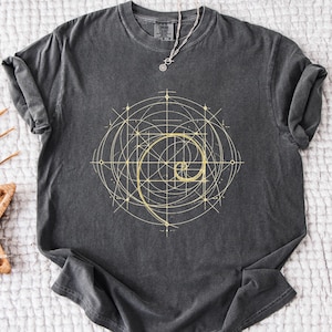 May include: Dark gray t-shirt featuring a golden spiral design with geometric lines. The shirt has a crew neck and short sleeves. A silver necklace with a pendant is draped on the shirt. The shirt is laying on a white surface.
