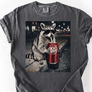 Może przedstawiać: Ciemnoszara koszulka z nadrukiem graficznym szopa pracza w okularach przeciwsłonecznych, pijącego z puszki Dr. Pepper. Szop siedzi na gzymsie z rozmytym tłem miejskim. Koszulka ma okrągły dekolt i krótkie rękawy.