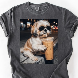 May include: Dark gray t-shirt featuring a graphic of a Shih Tzu dog wearing sunglasses and drinking iced coffee. The dog is brown and white. The image is set against a blurred background of city lights.