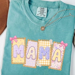 May include: Teal Comfort Colors t-shirt with the word "MAMA" in a colorful, patterned design. The letters are in individual frames with bows. A gold necklace with a pendant is on the shirt.