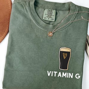 May include: An olive green t-shirt with a graphic of a pint of stout and the words "VITAMIN G" in white. A gold necklace with a small pendant is draped on the shirt. The shirt is folded, and a brown hat is partially visible.