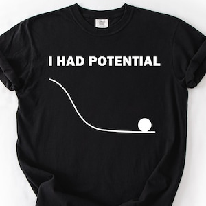 May include: Black t-shirt with the white text "I HAD POTENTIAL" above a white line drawing of a ball rolling down a curved path. The shirt is made of a soft material and has a crew neck.