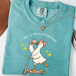 May include: Teal Comfort Colors t-shirt with a cartoon goose riding a pink scooter and drinking coffee. The text "Off to Regulate Emotions" is above the goose. A gold necklace is draped across the shirt.