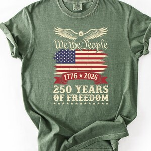 May include: A green t-shirt with a graphic design featuring the American flag, the text "We the People," and "250 Years of Freedom." The design includes the years 1776 and 2026, and a banner.