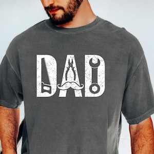 May include: Dark grey t-shirt featuring the word "DAD" in white. The letters are creatively designed with tools: a saw, pliers, and a wrench, with a moustache. A thoughtful gift for Father's Day.