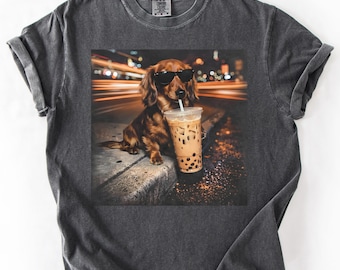 May include: Dark gray t-shirt featuring a graphic of a dachshund wearing sunglasses, holding a bubble tea. The dog is brown with black sunglasses. The background shows blurred city lights. The shirt is made of a soft material.