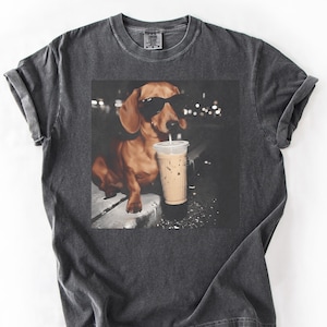 May include: Dark gray t-shirt featuring a graphic of a dachshund wearing sunglasses and drinking an iced coffee. The dog is brown with black sunglasses. The iced coffee is in a clear plastic cup with a white straw.