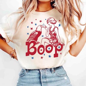 May include: A cream-colored t-shirt featuring a red and blue graphic print of a person touching the beak of a bald eagle. The word "Boop" is printed in large red letters below the figures, surrounded by small red and blue stars on the front.