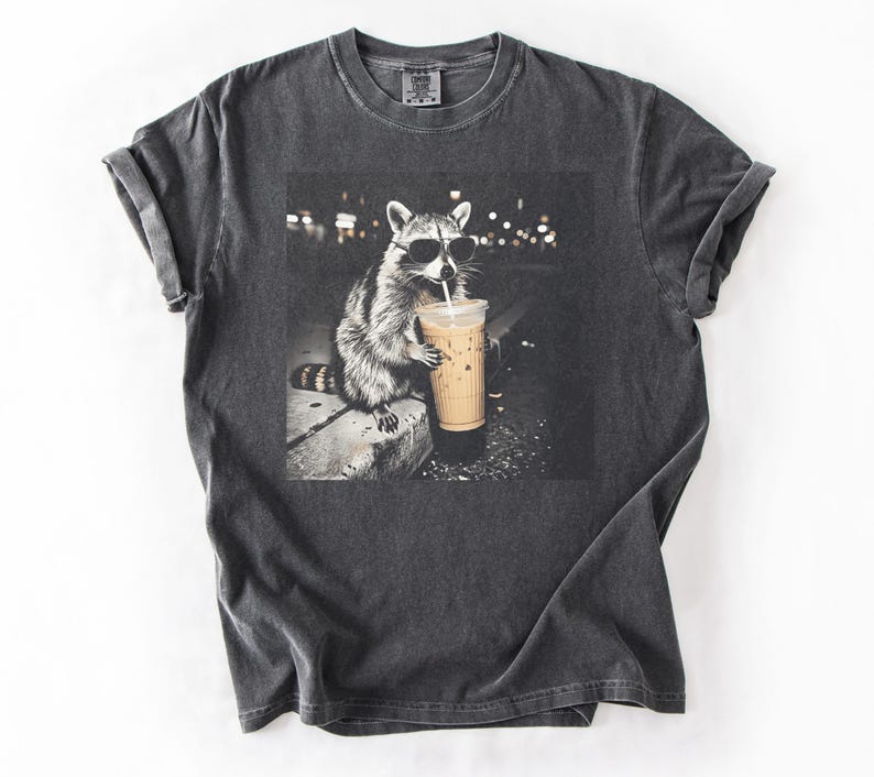 May include: Dark gray t-shirt featuring a graphic of a raccoon wearing sunglasses and drinking an iced coffee. The raccoon is in a black and white image, with a blurred background of city lights. The t-shirt is made of a soft, comfortable material.