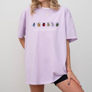 May include: A light purple t-shirt with the text "sad bug summer" and embroidered insect designs in various colors. The shirt is a relaxed fit and has short sleeves.