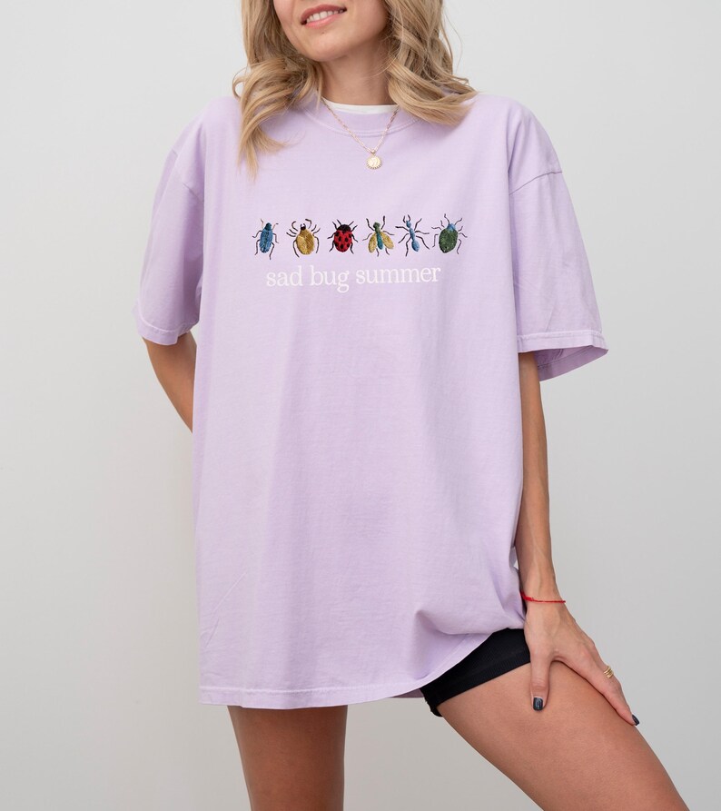May include: A light purple t-shirt with the text "sad bug summer" and embroidered insect designs in various colors. The shirt is a relaxed fit and has short sleeves.