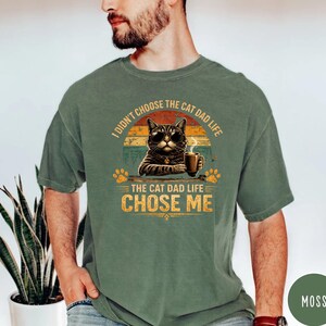 Op de afbeelding: Mosgroen T-shirt met een afbeelding van een kat met een zonnebril en een koffiekopje. De tekst luidt: "Ik heb niet het kattenvaderleven gekozen, het kattenvaderleven koos mij." Het ontwerp bevat pootafdrukken en een zonsondergang.