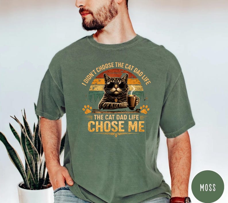 Op de afbeelding: Mosgroen T-shirt met een afbeelding van een kat met een zonnebril en een koffiekopje. De tekst luidt: "Ik heb niet het kattenvaderleven gekozen, het kattenvaderleven koos mij." Het ontwerp bevat pootafdrukken en een zonsondergang.