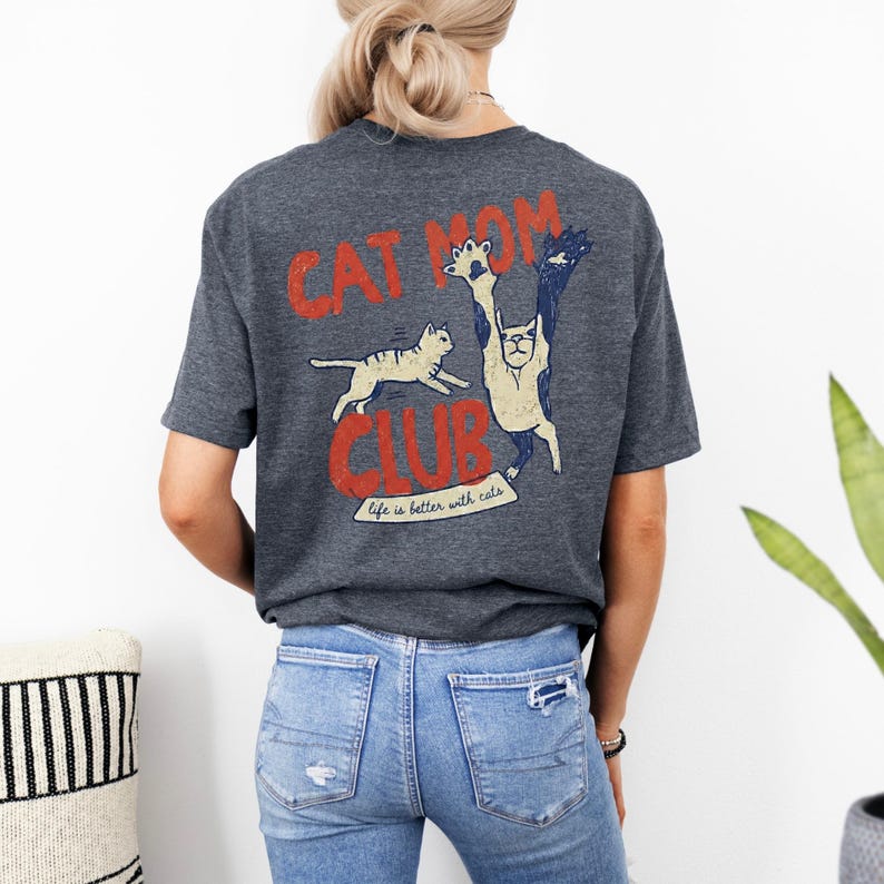 May include: Dark gray t-shirt with a distressed red and blue graphic that reads "CAT MOM CLUB." The graphic features cartoon cats in various poses, with the phrase "Life is better with cats" below. The shirt is worn by a person.