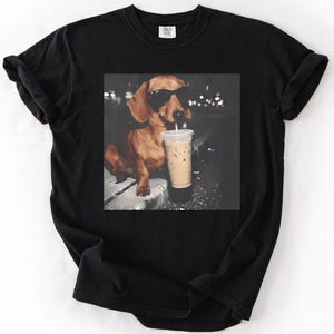 May include: Black t-shirt featuring a graphic of a dachshund wearing sunglasses and drinking an iced coffee. The dog is brown with black sunglasses and a white straw in the coffee cup. The background is dark with blurred lights.