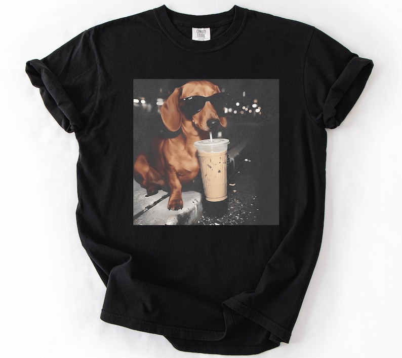 May include: Black t-shirt featuring a graphic of a dachshund wearing sunglasses and drinking an iced coffee. The dog is brown with black sunglasses and a white straw in the coffee cup. The background is dark with blurred lights.
