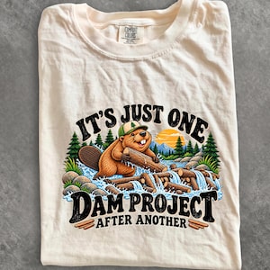May include: A cream-coloured t-shirt featuring a graphic of a beaver wearing a green and white cap, holding a log in a river with a dam. The text reads IT'S JUST ONE DAM PROJECT AFTER ANOTHER. The shirt has a Comfort Colors brand label at the neck.