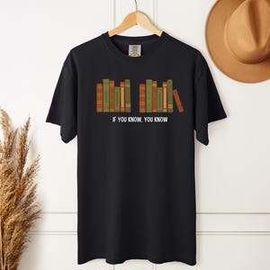 May include: Black t-shirt with a graphic of a row of books in shades of green, orange, and brown. The text "IF YOU KNOW, YOU KNOW" is printed in white below the books. The shirt is hanging on a wooden hanger.