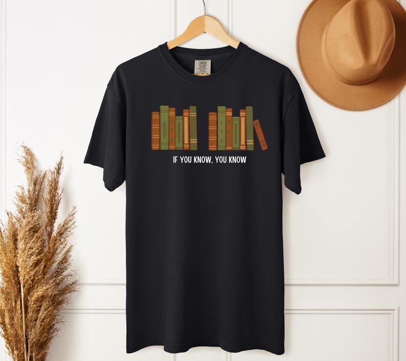 May include: Black t-shirt with a graphic of a row of books in shades of green, orange, and brown. The text "IF YOU KNOW, YOU KNOW" is printed in white below the books. The shirt is hanging on a wooden hanger.