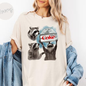 May include: Ivory-colored t-shirt featuring a graphic of three raccoons. One raccoon is holding a Diet Coke can in front of a moon, while the others look on. The text "Diet Coke" is visible on the can.
